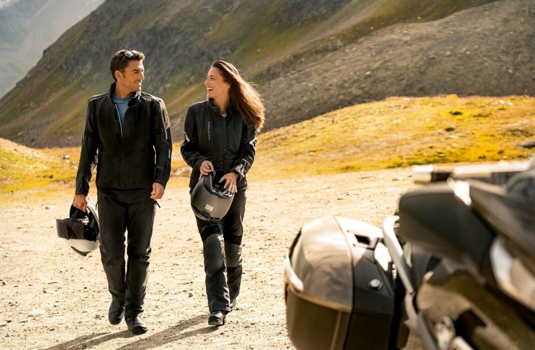 Mann und Frau mit Helm in der Hand lachend in Motorradkleidung: Leder oder Textil auswählen